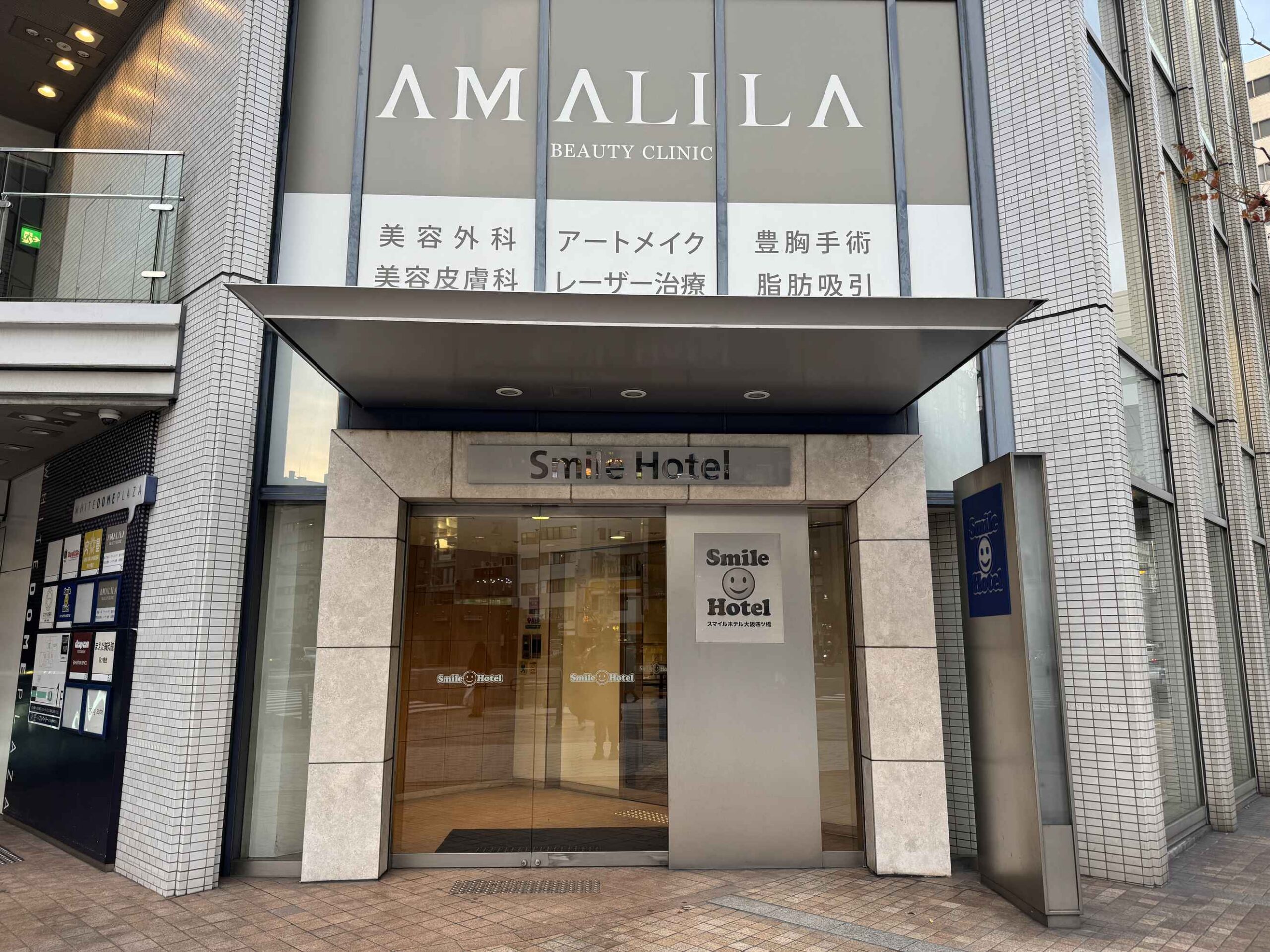大阪微笑飯店四ツ橋分店入口外觀，玻璃門上方Smile Hotel黃色招牌與AMALILA美容診所共用大樓