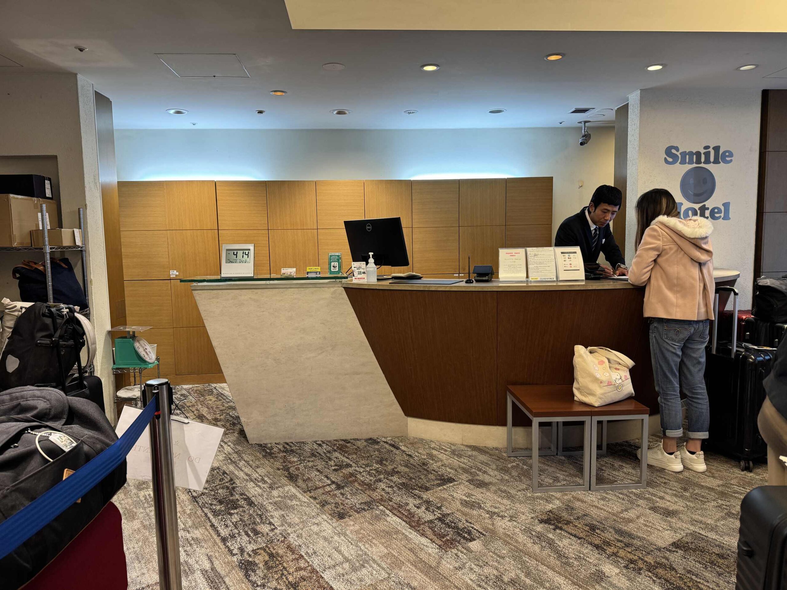 大阪微笑飯店一樓櫃檯，飯店人員正幫旅客辦理入住，後方是Smile Hotel品牌牆面與藍色logo