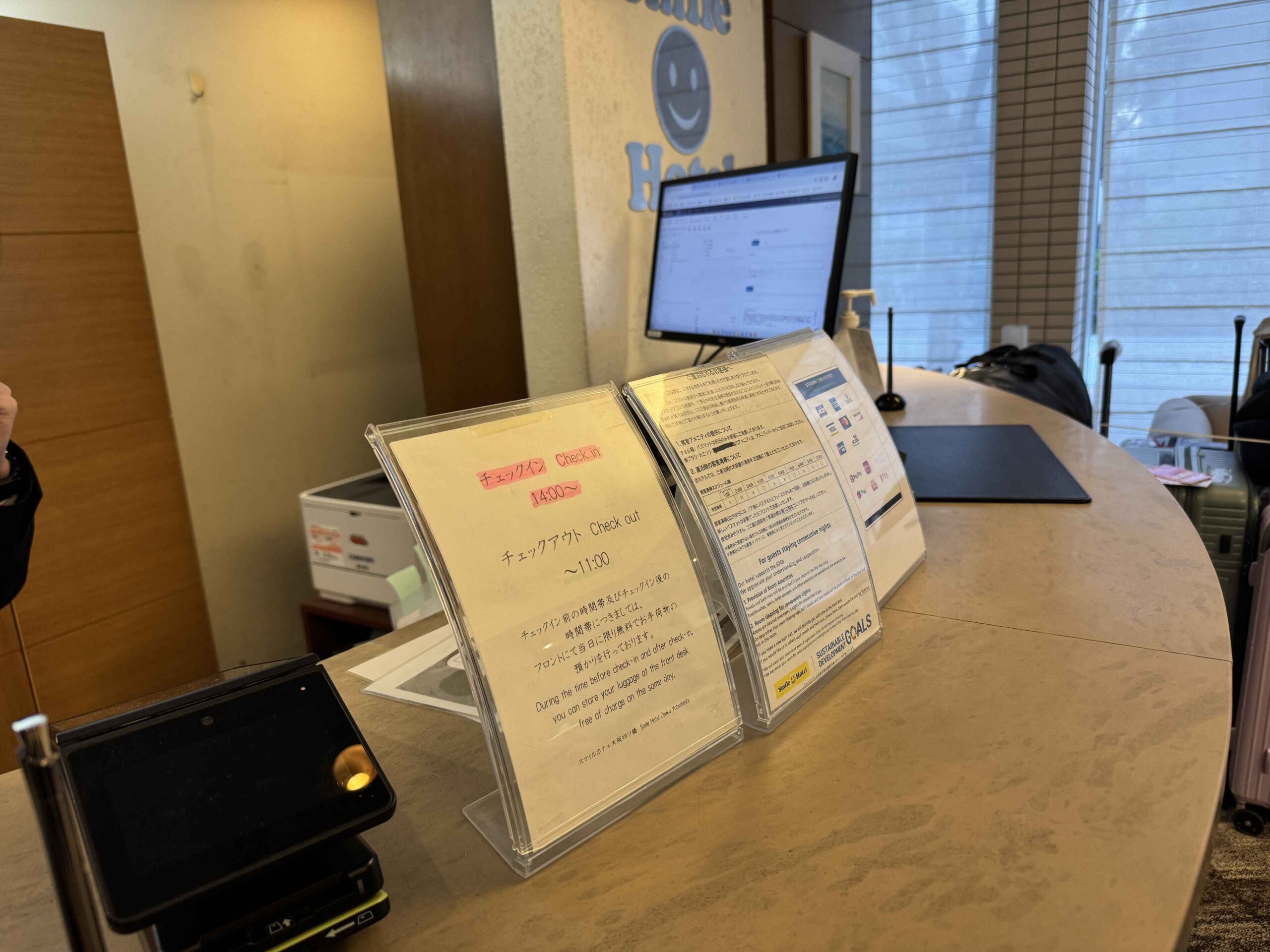 大阪微笑飯店櫃檯桌上的check-in時間告示卡，標示14:00入住、11:00退房，並提供當日免費行李寄放