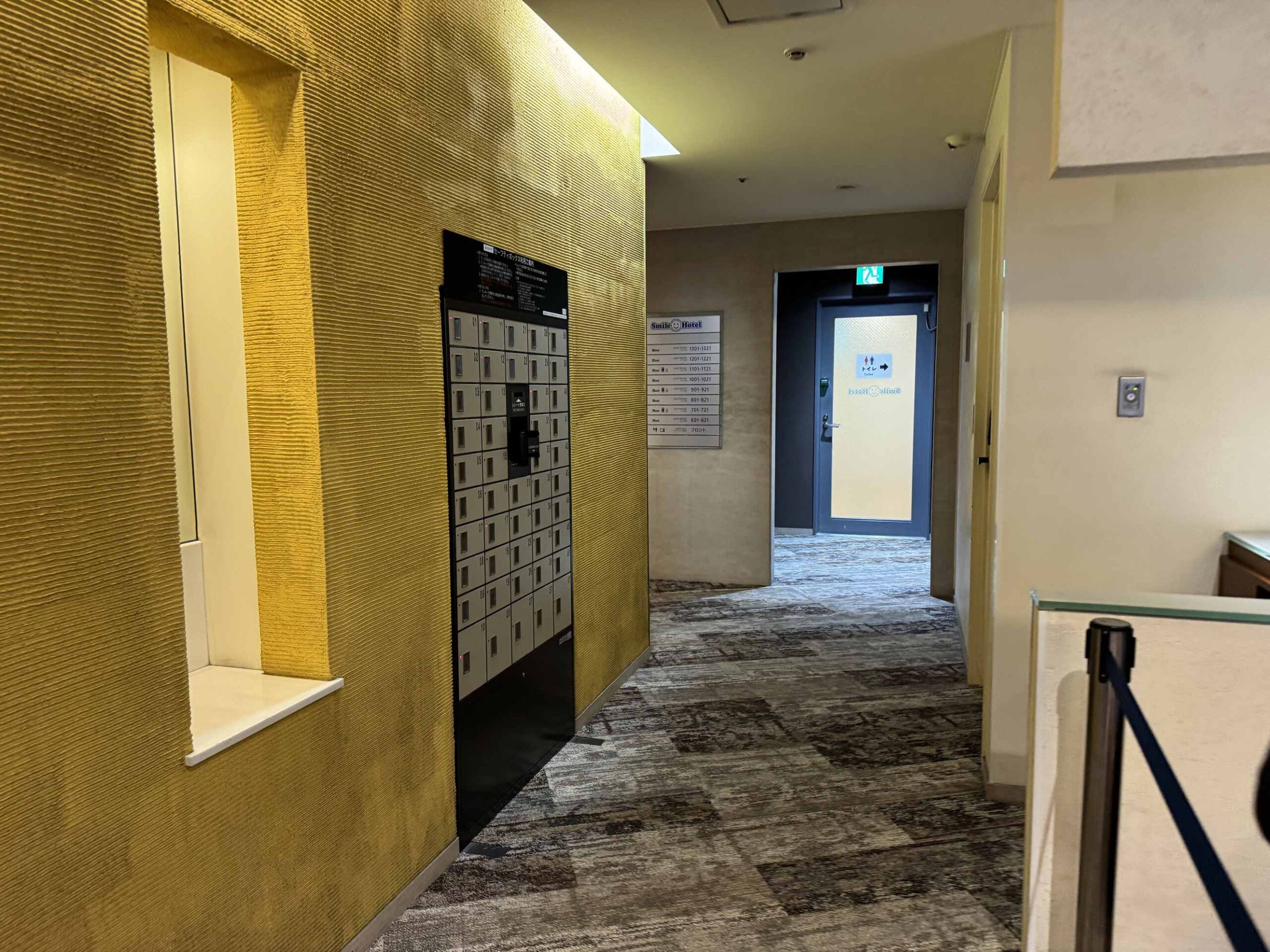 大阪微笑飯店電梯廳走廊，金色編織紋牆面搭配貴重物品保險箱櫃與公共廁所指示牌
