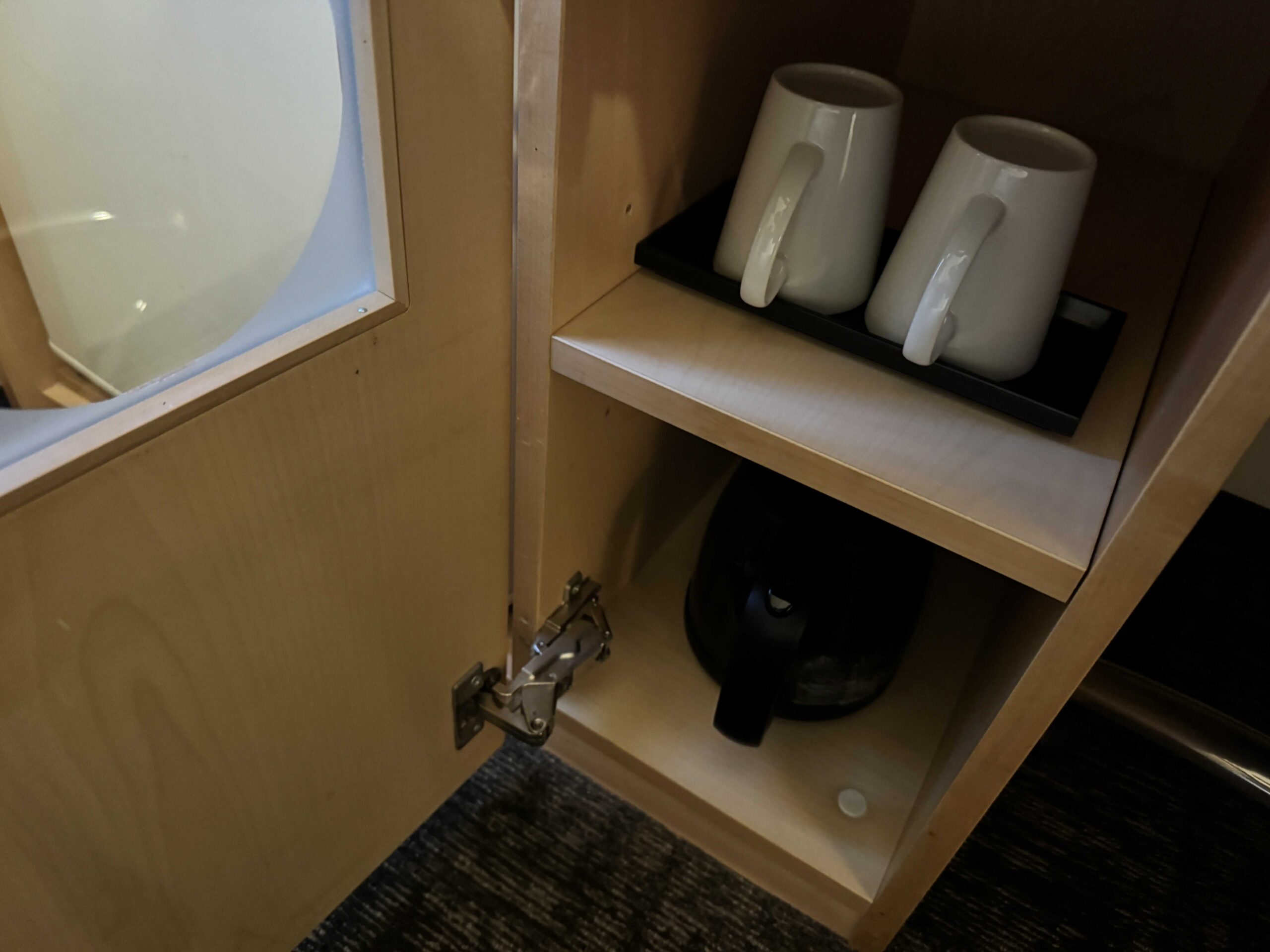 飯店茶水區木製收納櫃，上層放置兩個白瓷馬克杯、下層擺有黑色快煮壺與杯墊