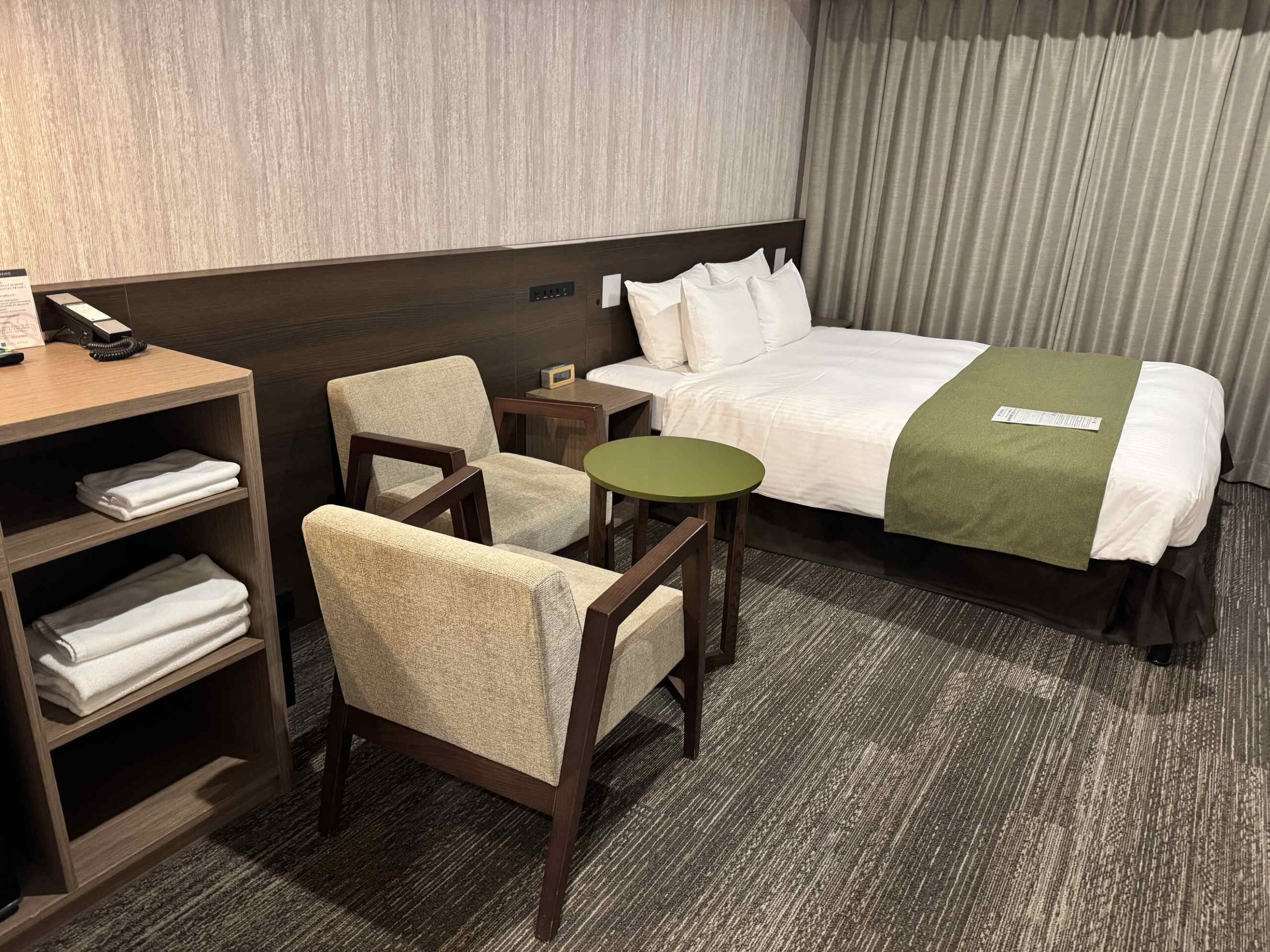KOKO HOTEL京都三條標準雙人房主視角，雙人床配軍綠色腳尾巾、沙發椅與綠色小圓桌、靠牆書桌與毛巾收納櫃
